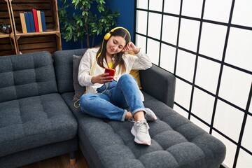 Young hispanic woman listening to music sitting on sofa at home