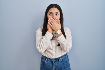 Young latin woman standing over blue background shocked covering mouth with hands for mistake. secret concept.