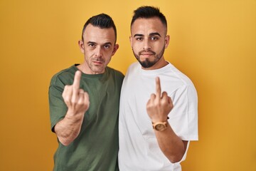 Homosexual couple standing over yellow background showing middle finger, impolite and rude fuck off expression