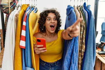 Young hispanic woman searching clothes on clothing rack using smartphone pointing with finger surprised ahead, open mouth amazed expression, something on the front