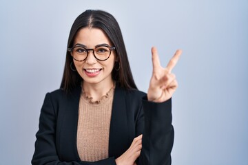 Young brunette woman standing over blue background smiling with happy face winking at the camera doing victory sign. number two.