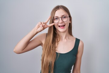 Young caucasian woman standing over white background doing peace symbol with fingers over face, smiling cheerful showing victory