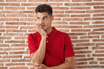 Young hispanic man standing over bricks wall looking stressed and nervous with hands on mouth biting nails. anxiety problem.