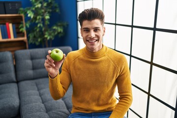 Young hispanic man holding green apple looking positive and happy standing and smiling with a confident smile showing teeth