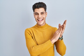 Young hispanic man standing over blue background clapping and applauding happy and joyful, smiling...