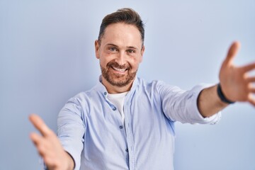 Fototapeta premium Middle age caucasian man standing over blue background looking at the camera smiling with open arms for hug. cheerful expression embracing happiness.
