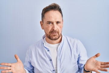 Middle age caucasian man standing over blue background clueless and confused with open arms, no idea concept.