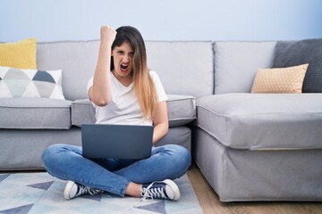Obraz premium Young brunette woman sitting on the floor at home using laptop annoyed and frustrated shouting with anger, yelling crazy with anger and hand raised
