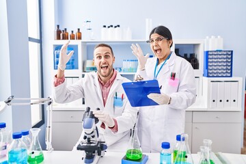 Two young people working at scientist laboratory celebrating victory with happy smile and winner expression with raised hands