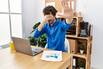 Young hispanic man with beard working at the office using computer laptop covering eyes with hands and doing stop gesture with sad and fear expression. embarrassed and negative concept.