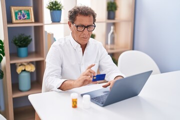 Middle age man using laptop and credit card sitting on table with pills bottles at home