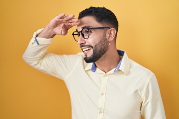 Fototapeta premium Hispanic young man wearing business clothes and glasses very happy and smiling looking far away with hand over head. searching concept.