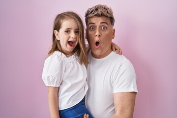 Young father hugging daughter over pink background scared and amazed with open mouth for surprise, disbelief face