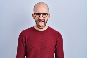 Young bald man with beard standing over white background wearing glasses sticking tongue out happy with funny expression. emotion concept.