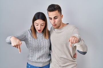 Young hispanic couple standing over white background pointing down with fingers showing advertisement, surprised face and open mouth