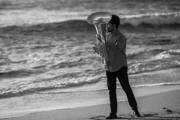 A man musician playing a tuba on the ocean beach. Black and white photo.