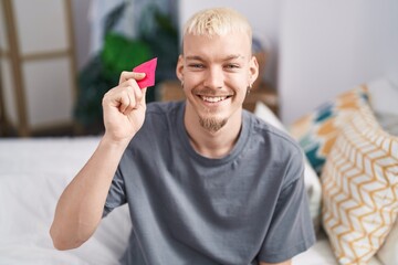 Young caucasian man holding condom sitting on bed looking positive and happy standing and smiling with a confident smile showing teeth - obrazy, fototapety, plakaty