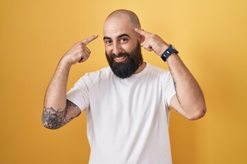 Young hispanic man with beard and tattoos standing over yellow background smiling pointing to head...