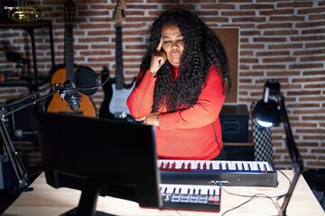 Plus size hispanic woman playing piano at music studio with hand on chin thinking about question, pensive expression. smiling with thoughtful face. doubt concept.