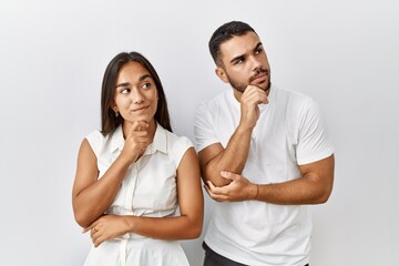 Fototapeta premium Young interracial couple standing together in love over isolated background with hand on chin thinking about question, pensive expression. smiling with thoughtful face. doubt concept.