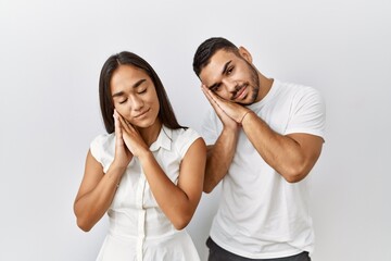 Young interracial couple standing together in love over isolated background sleeping tired dreaming and posing with hands together while smiling with closed eyes.