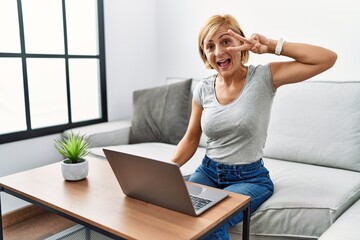 Middle age blonde woman using laptop at home doing peace symbol with fingers over face, smiling...