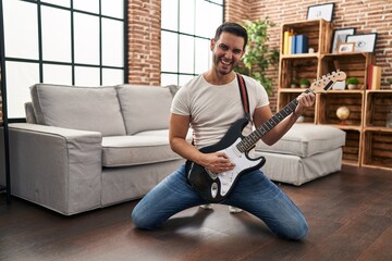 Young hispanic man playing electrical guitar with knees on floor at home