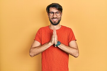 Young hispanic man wearing casual clothes and glasses praying with hands together asking for forgiveness smiling confident.