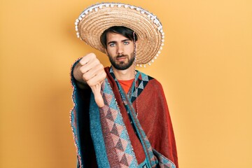 Young hispanic man holding mexican hat looking unhappy and angry showing rejection and negative with thumbs down gesture. bad expression.