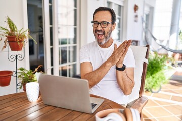 Middle age man using computer laptop at home clapping and applauding happy and joyful, smiling proud hands together