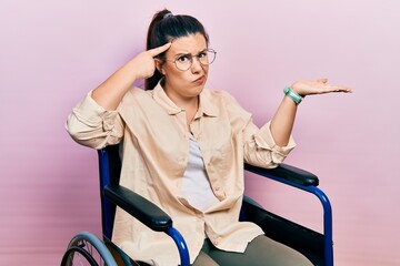 Young hispanic woman sitting on wheelchair confused and annoyed with open palm showing copy space...