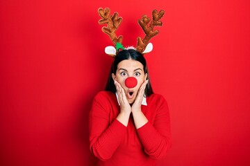 Young hispanic woman wearing deer christmas hat and red nose afraid and shocked, surprise and...