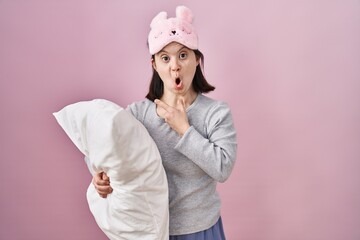 Woman with down syndrome wearing sleeping mask hugging pillow surprised pointing with finger to the side, open mouth amazed expression.