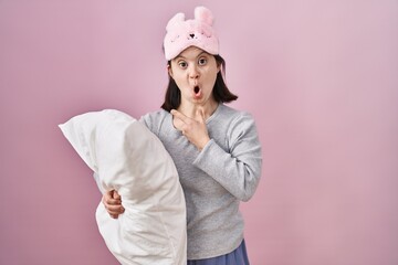 Woman with down syndrome wearing sleeping mask hugging pillow surprised pointing with finger to the...