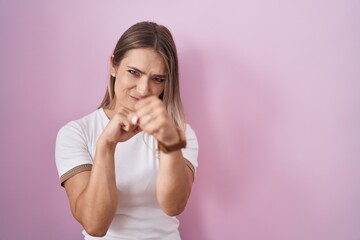 Blonde caucasian woman standing over pink background punching fist to fight, aggressive and angry attack, threat and violence