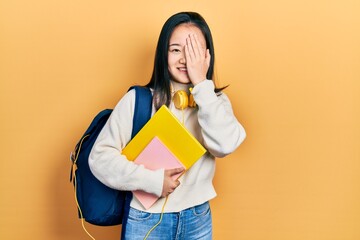 Young chinese girl holding student backpack and books covering one eye with hand, confident smile on face and surprise emotion.