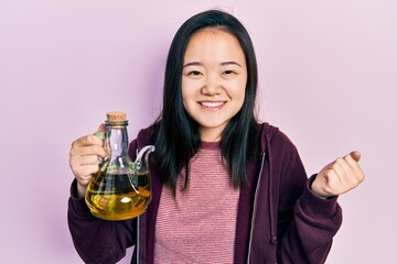 Young chinese girl holding olive oil can screaming proud, celebrating victory and success very...