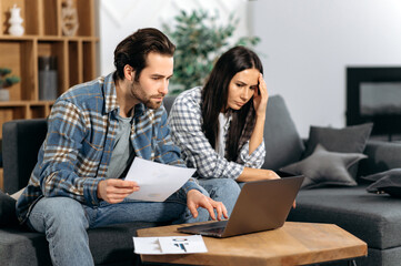 Frustrated caucasian couple sitting on the couch, the guy and the girl are freelancers, work from home, use a laptop, look at graphs, are unhappy with the results, low profit, thinking about strategy