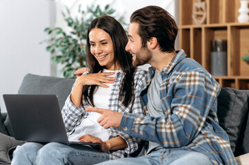 Positive spouses spending time together at home, they sitting on comfortable sofa in the living room, use a laptop, make online purchases, watch news, browse social networks, smile together