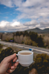 Hand holding a hot latte coffee in a white cup over a blurry background of a landscape with forest, mountains, blue sky and a highway