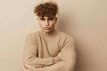 a close horizontal portrait of a handsome, charming man with curly hair standing in a beige sweater on a beige background, hugging himself with his arms