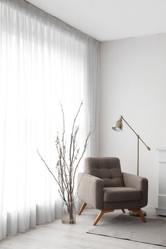 Vase with tree branches, armchair and lamp near light curtain in living room