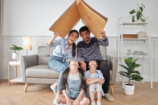Asian Thai family, adult dad, mum, and little children happily fun playing in a white living room, built cardboard paper safety house with imagination, lovely weekend and wellbeing domestic lifestyle.