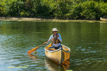 A woman of retirement age solo paddles a canoe around the Toronto Islands wearing sun safe clothing...