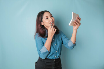 A portrait of a beautiful young Asian woman applying lipstick on her lips, isolated by a blue...