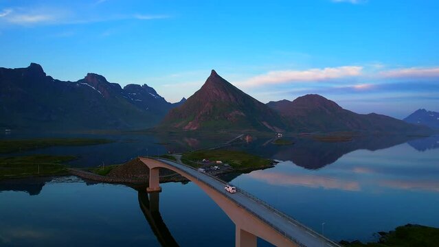 Aerial following shot of a converted van driving over famous bridges Fredvang on Lofoten Islands with Volandstind reflecting on the water