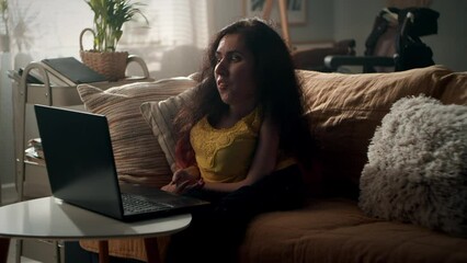 Woman with disability talking via video call with friend using laptop while sitting on a couch in cozy living room in her spare time