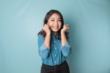 A portrait of a cute Asian woman wearing a blue shirt and feeling excited and isolated by a blue background