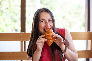 Young beautiful woman in red blouse eating croissant at a cafe. Hungry female having breakfast out while eating tasty croissant with pleasure.