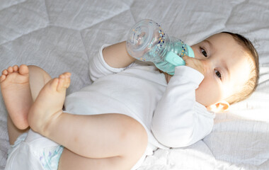 baby lies on bed using plastic water bottle.toddler playing holding bottle over the head chewing silicone head.bedroom interior sunny morning white blanket.kid hydration concept healthy care love 